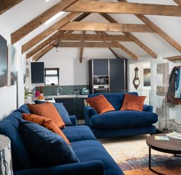 A living room with blue sofas and orange cushions with a round wooden coffee table and an open plan kitchen under wooden ceiling beams at The Rum House in Porthallow