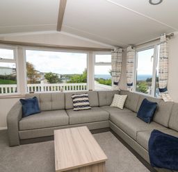 A living room with a gray sectional sofa and cushions with a wooden coffee table and windows showing outdoor greenery at Gwent 74 in Borth
