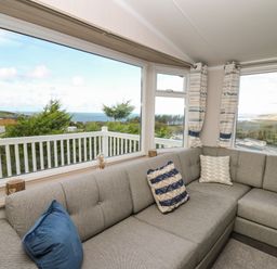 A living room with a gray sectional sofa and blue and white cushions by large windows overlooking greenery and a seaside view at Gwent 74 in Borth