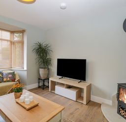 A living room with a TV on a stand a wood burning stove a wooden coffee table and a sofa with floral cushions at Birch House in Corsham