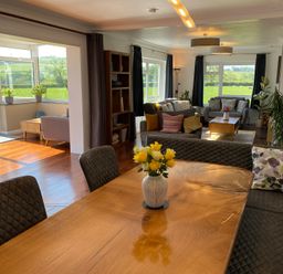 A dining table with a flower vase and cushioned chairs next to a living area with sofas and large windows at Ashlea in Launceston
