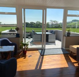 A sunlit living room with blue chairs plants and a wooden table overlooking a patio with outdoor furniture and green fields at Ashlea Launceston