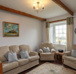 A living room with beige sofas cushions a small wooden table a window with curtains a wall clock and a framed painting at No2 Budle Bay Cottage Bamburgh