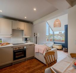 A kitchen with cabinets and an oven next to a dining table and a living area with a sofa and a large window at Castle View Bamburgh