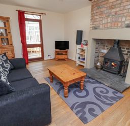 A living room with a black sofa and patterned cushions a wooden coffee table on a patterned rug a wood burning stove in a brick fireplace a wooden cabinet a television on a wooden stand a white shelving unit and a door with a red curtain at St Marys Cottages No1 in Low Newton-by-the-Sea