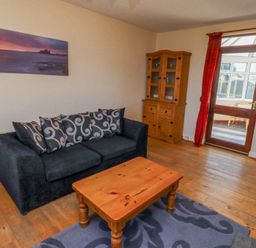 A living room with a black sofa with patterned cushions a wooden coffee table a wooden cabinet and a door leading to a conservatory at St Marys Cottages No1 in Low Newton-by-the-Sea