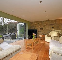 A living room with beige sofas wooden tables and a television with an open door leading to an outdoor patio at Quarry Haven in Bamburgh