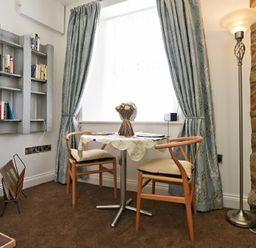 A small dining area with two chairs a round table with a tablecloth and flowers by a window with curtains and a floor lamp near a stone wall at Sea Air in Amble