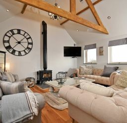 A living room with beige and gray sofas a wooden coffee table a black wood stove a large wall clock and exposed wooden ceiling beams at The Hemmel (Beadnell) in Beadnell