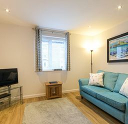 A living room with a teal sofa and white cushions a floor lamp a framed beach painting a window with curtains a small wooden table a tv on a glass stand and wooden cabinet at Braidcarr Cottage in Seahouses