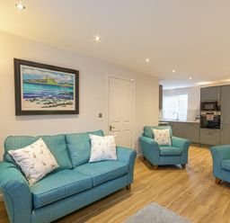 A living room with blue sofas and armchairs and a kitchen area with grey cabinets in the background at Braidcarr Cottage Seahouses