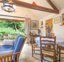 A dining area with wooden chairs and a table with a vase of flowers and a blue sofa next to large open doors showing a garden at The Byre at Warkworth
