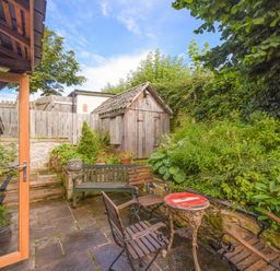An outdoor patio area with wooden chairs a small round table a wooden bench stone steps greenery and a small wooden shed at The Byre at Warkworth in Warkworth