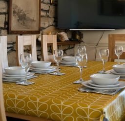 A dining table with plates bowls and wine glasses set on a yellow patterned tablecloth in a room with a stone wall and wooden chairs at Lobster Lodge in Seahouses