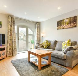 A living room with a grey sofa, wooden coffee table, television on a wooden stand and glass door with floral curtains at East Havers in Alnwick