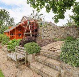 A garden with a wooden bench a children's swing set a wooden playhouse stone steps and a stone wall at Manor Cottage in Embleton
