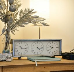 A wooden table with a clock showing three time zones a glass vase with decorative branches a notebook and a black device with a dock at West End Cottage in Embleton