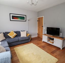 A living room with a gray sofa, yellow and beige cushions, a yellow rug, a wooden TV stand with a TV, a wooden door, and a floor lamp at Alnside Cottage in Alnmouth
