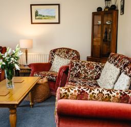 A living room with floral patterned armchairs and sofa a wooden coffee table with flowers a lamp and a display cabinet at Swallows Cottage nr Warkworth in Acklington