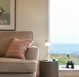 A sofa with a patterned cushion next to a small table with a lamp and a window showing a sea view at Portside in Seahouses