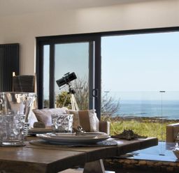 A dining area with a wooden table set with plates and glasses and a telescope by sliding glass doors overlooking the sea at Waters Edge Beadnell Harbour Road in Beadnell