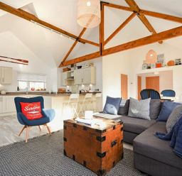 An open living space with a gray sectional sofa a wooden trunk coffee table a blue armchair with a red cushion a kitchen with bar stools and exposed wooden ceiling beams at Bowline Cottage in Beadnell