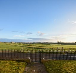 A fenced grassy garden area with a paved path overlooking a green field under a blue sky at Mariners House in Alnmouth