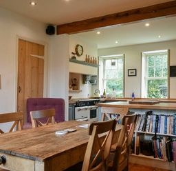 A kitchen with wooden dining table and chairs wooden countertop and bookshelf beneath windows at Whistlewood in Bamburgh