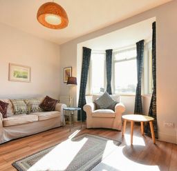 A living room with a sofa, armchair, lamp, two small tables, rug, TV, and bay windows with dark curtains at Rosedale House in Amble