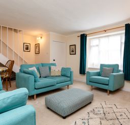 A living room with turquoise sofas a patterned ottoman curtains and a dining table under stairs at Percy Cottages No4 in Alnmouth