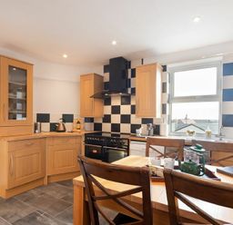 A kitchen with wooden cabinets a black and white checkered backsplash a black stove a wooden dining table with chairs and a window at Church View in Bamburgh