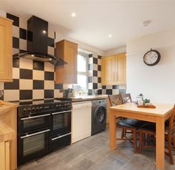A kitchen with wooden cabinets, a black stove, checkered black and white backsplash, a wooden dining table with chairs and a clock on the wall at Church View in Bamburgh