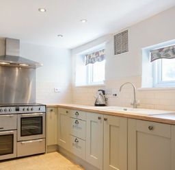 A kitchen with a stainless steel stove and hood a kettle on the countertop and twin windows with floral blinds at Cargoes in Craster