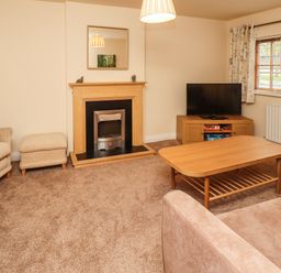 A living room with a beige armchair, ottoman, coffee table, TV on a wooden cabinet, fireplace, and windows with curtains at Stewart House in Wooler