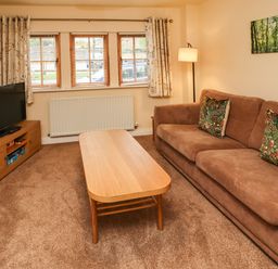 A living room with a brown sofa and patterned cushions a wooden coffee table a television on a wooden stand and a floor lamp at Stewart House in Wooler