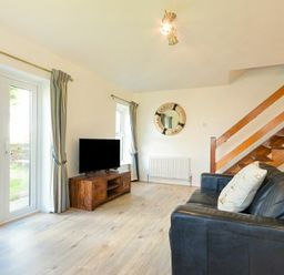 A living room with a black leather sofa a wooden TV stand with a TV curtains on windows and a wooden staircase at Coastal Plaice in Beadnell
