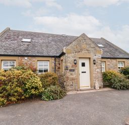 Stone house with white door and multiple windows surrounded by bushes at The Wynd in Bamburgh