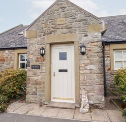 Stone house exterior with a white door and two outdoor wall lights plants on either side of entrance at The Wynd in Bamburgh