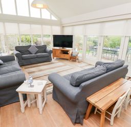 A living room with three blue sofas a wooden table with small chairs a television and large windows at The Wynd in Bamburgh