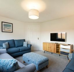 A living room with blue sofas a patterned ottoman a wooden TV stand and a framed picture on the wall at Luna Tide in Seahouses