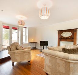 A living room with cream sofas a wooden floor a small TV an ornate wooden fireplace and large windows at The Old Concert Room in Alnmouth