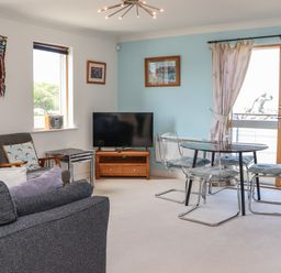 A living room with a gray sofa a glass dining table with four chairs a television on a wooden stand and large windows at Castle View at the Viking in Seahouses