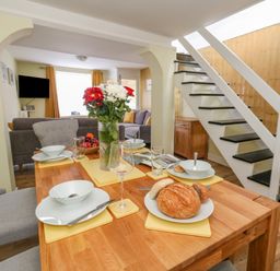A dining table set with plates bowls glasses and bread next to stairs and a living room at Cove Street Cottage in Weymouth