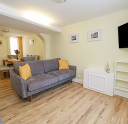 A living room with a gray sofa with yellow pillows a wall-mounted TV and a bookshelf with books and a dining area in the background at Cove Street Cottage Weymouth