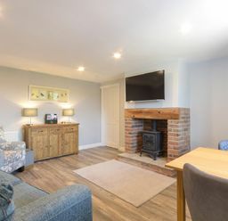 A living room with a brick fireplace and TV above it a wooden sideboard with lamps a floral armchair and a wooden dining table with chairs at Dairymaid's Cottage in Amble