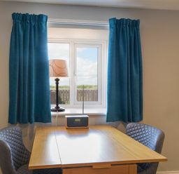 A small wooden table between two gray chairs in front of a window with blue curtains and a lamp and radio on the windowsill at Dairymaid's Cottage in Amble