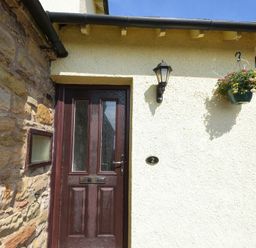 A wooden front door with glass panels on a stone and white wall exterior with a wall lamp and hanging flower pot at Dairyman's Cottage in Amble