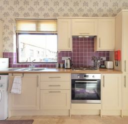 A kitchen with cream cabinets purple tile backsplash and a washing machine under the counter at Dairyman's Cottage in Amble