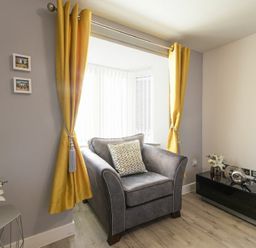 A sitting area with a gray armchair yellow curtains side table and black cabinet with decorations