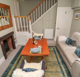 A living room with a beige sofa a wooden table set for two and a staircase at Five Bells Cottage in Alnmouth